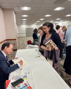 Behind the Books Chris Bohjalian (2) A man in a suit sits at a table signing a book for a woman holding several books. Other people wait in line, and the scene takes place in a brightly lit, carpeted room with white walls.