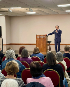 Behind the Books Chris Bohjalian A man in a navy suit speaks at a podium labeled ALEPPO to an audience seated in red chairs in a conference room with beige walls and ceiling lights.