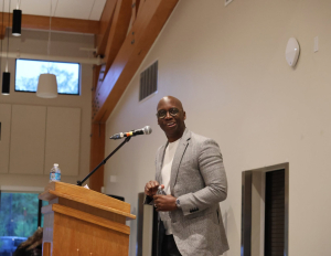 Behind the Books Michael Holley (2) A man in a gray blazer and glasses stands at a podium with a microphone, smiling, in a well-lit room with wooden beams and large windows. A water bottle sits on the podium.