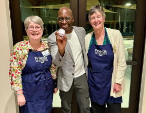Behind the Books Michael Holley Three people smile at the camera indoors. The two women wear navy Friends aprons and stand on either side of a man in a gray blazer who holds a signed baseball. All appear happy and are posing together.