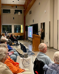 Behind the Books Sy Montgomery (2) A woman stands at a podium giving a presentation to an audience seated in a well-lit room. A screen beside her displays a photo, and people are attentively listening to the speaker.