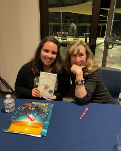 Behind the Books Sy Montgomery Two women sit at a table indoors. One woman smiles and holds up the book Soul of an Octopus, while the other woman leans on her hand and smiles. On the table are a notebook with a colorful cover, a pen, and a water bottle.