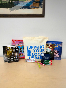 A tote bag reading Support Your Local Library sits on a table with books, pencils, a Friends-themed notepad, a sticker, and a Pet Adoption kit in front of a painting on the wall.
