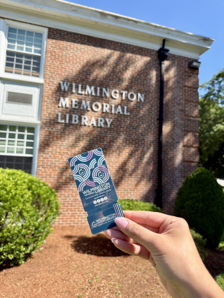 A hand holds a Wilmington Memorial Library card outside the library building, with the buildings name visible on the brick wall in the background.