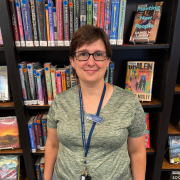 A person wearing glasses and a green shirt stands in front of library bookshelves, smiling. They have short brown hair and a blue lanyard with a name badge and keys. Books are visible in the background.