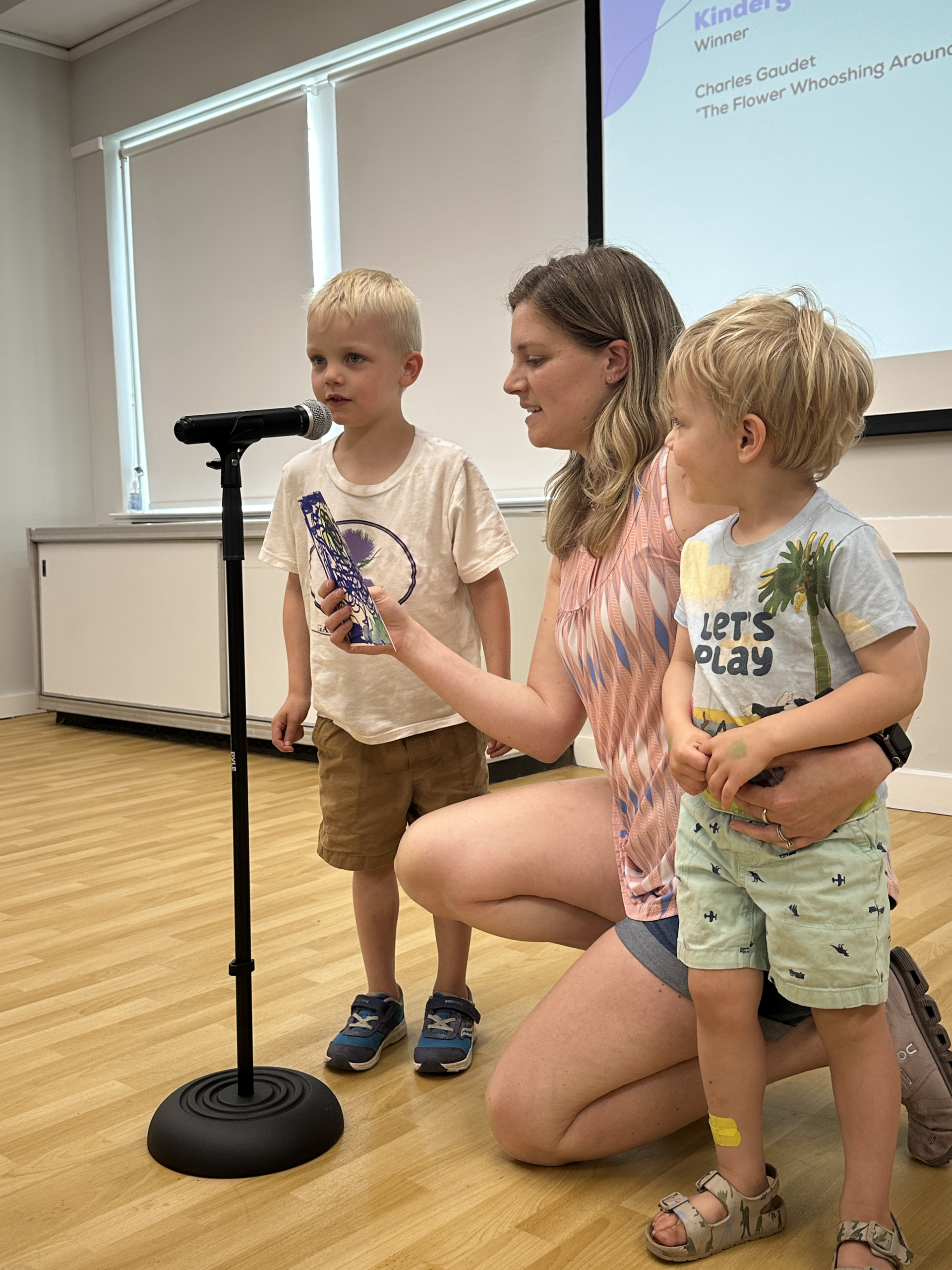 A woman kneels and holds a microphone and a decorated tube for a young boy speaking, while another toddler stands beside her. They are in a bright room with a screen and presentation in the background.