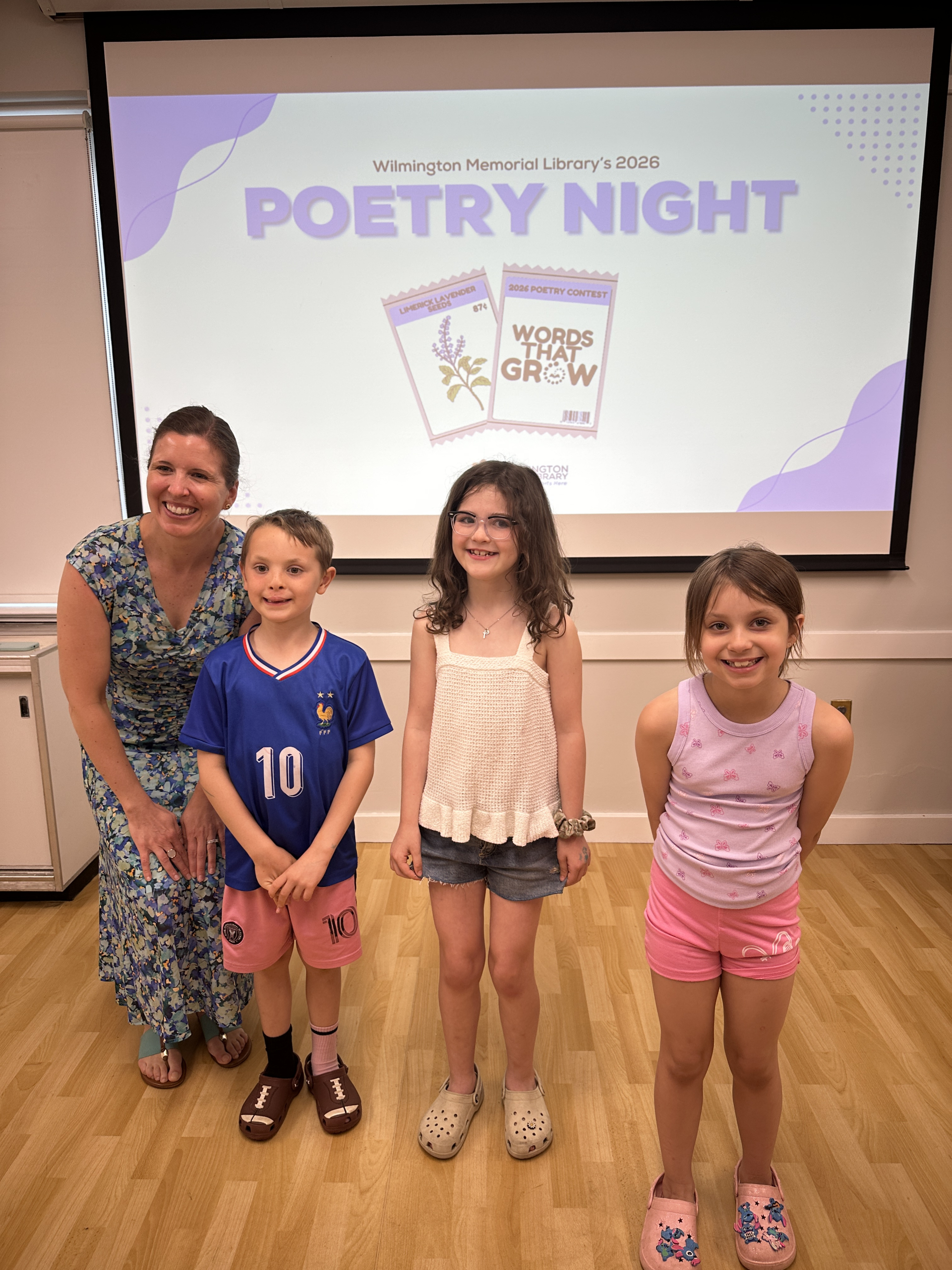 A smiling woman and three children pose together indoors in front of a projector screen displaying “Wilmington Memorial Library’s 2024 Poetry Night” and an image of a book titled “Words Grow.”.