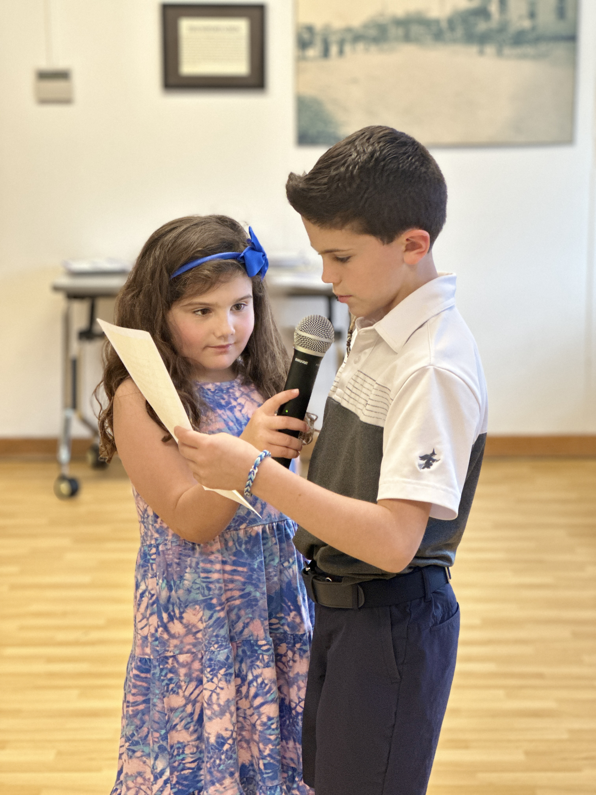 Two children stand indoors; the boy holds a microphone and a piece of paper as the girl looks on attentively. Both are dressed nicely, with the girl wearing a blue dress and bow, and the boy in a collared shirt and navy pants.