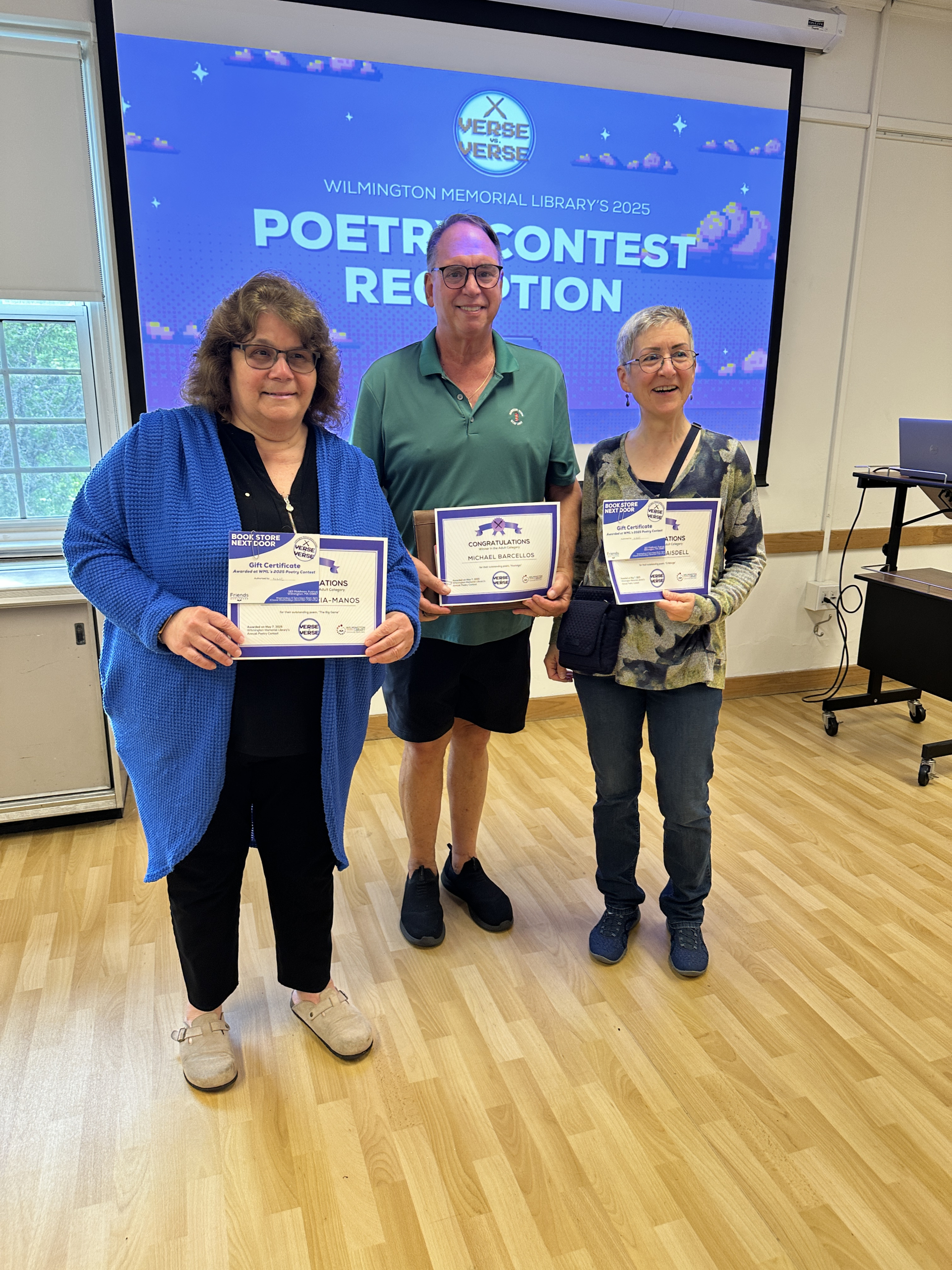 Poetry Night (6) Three adults stand indoors, holding certificates and smiling at the camera. Behind them is a screen displaying Wilmington Memorial Librarys 2025 Poetry Contest Reception with the Verse Versus logo at the top.