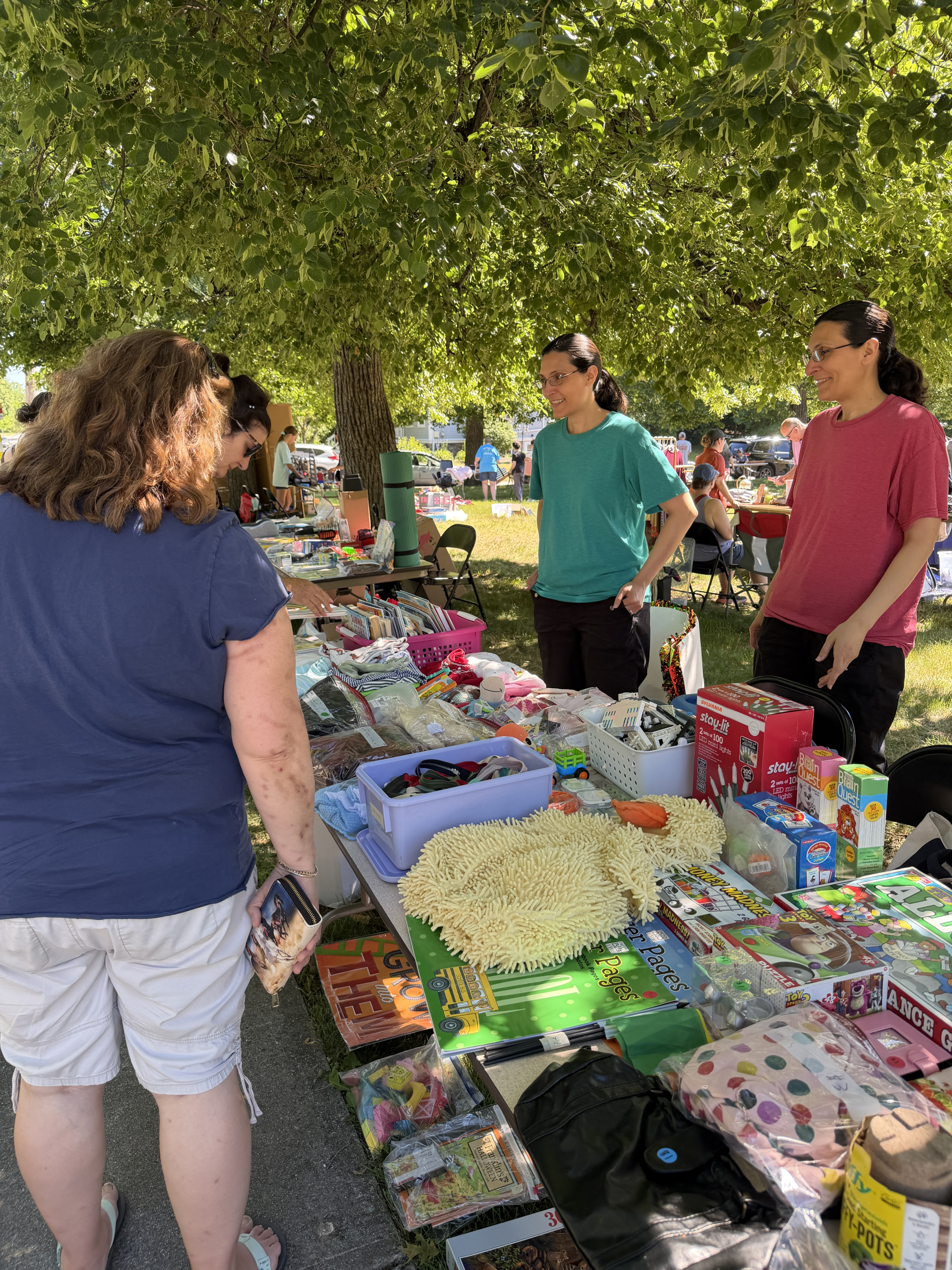 Three people stand around a table at an outdoor market, which is shaded by trees and covered with various toys, games, and household items. One person browses while two others talk and watch.
