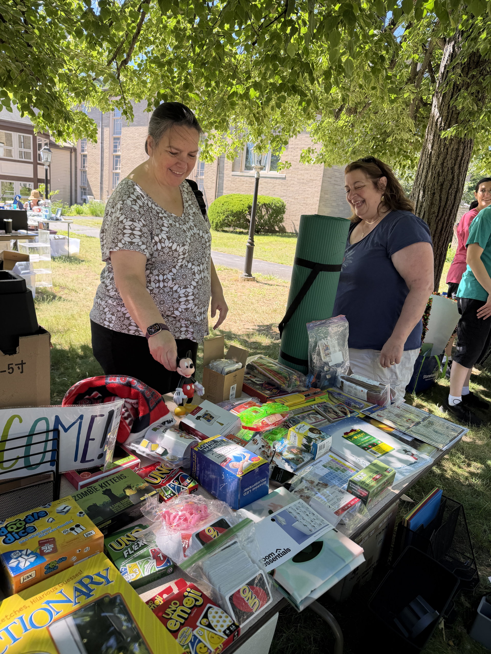 Two women smile while browsing and selling board games, toys, and various items at an outdoor table under leafy trees. The table is filled with games and colorful objects, and a “WELCOME!” sign is visible.