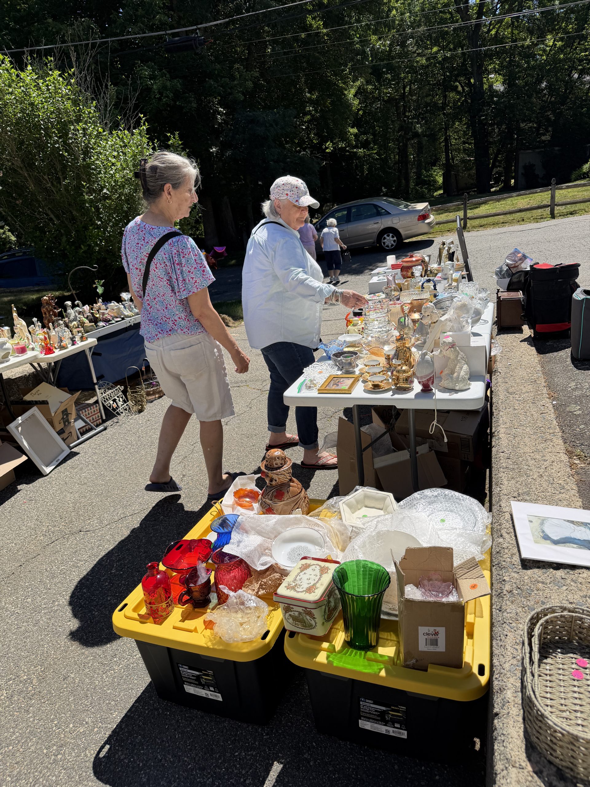 Two women browse tables full of vintage glassware, ceramics, and household items at an outdoor flea market on a sunny day. Various colorful items are displayed on bins and tables along the street.