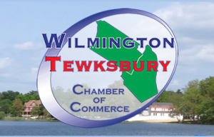 A slanted blue circle with the shapes of the towns of Wilmington and Tewksbury in green and the words Wilmington (in all caps and in blue font) and the word Tewsbury (in all caps in red font) over the town maps. Under that, but still in the circle are the words Chamber Of Commerce, with each word in caps below the one before it.